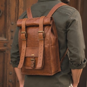 Può includere: Uno zaino in pelle marrone con chiusura a rullo, dotato di due cinghie verticali con fibbie in metallo color ottone, una tasca frontale con chiusura a fibbia singola e spallacci regolabili. La borsa ha una finitura liscia e una forma rettangolare.
