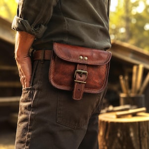 May include: A brown leather belt pouch featuring a rectangular flap with two brass-colored rivets and a central buckle closure. The pouch is attached to a matching brown leather belt worn around the waist, showcasing visible stitching along the edges.