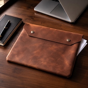 May include: A brown leather document sleeve featuring a fold-over flap secured by two metallic snap buttons. The rectangular case has a smooth, matte texture and contains white paper sheets visible at the side, resting on a dark wooden surface.