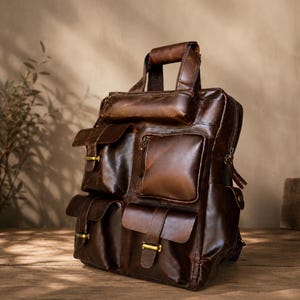 May include: A dark brown leather backpack featuring a top handle, multiple exterior pockets with flap closures, and brass-toned hardware. The bag has a structured design with visible stitching and a smooth, polished finish on the leather surface.