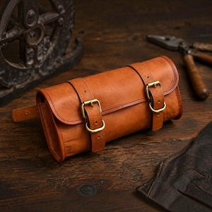 Könnte beinhalten: Eine zylindrische Werkzeugrolle aus hellbraunem Leder mit zwei vertikalen Riemen und messingfarbenen Metallschnallen. Das Leder hat eine glatte, matte Oberfläche mit sichtbaren Nähten entlang der Riemenkanten und am Hauptkörper des Behälters.