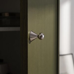 May include: A polished silver-toned metal cabinet knob featuring a conical base that tapers towards a spherical head. The knob is mounted on a vertical, flat, olive-green panel with a subtle brushed texture, showing a clean and reflective surface.