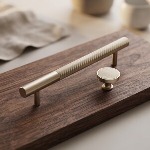 Op de afbeelding: Een kastgreep van geborsteld goud en een bijpassende ronde kastknop. De greep heeft een lange, cilindrische stang met een textuurrijk, geribbeld middengedeelte en gladde uiteinden. De ronde knop heeft een plat, geribbeld bovenoppervlak.