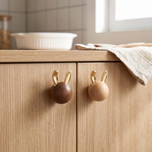 Puede incluir: Dos pomos de armario esféricos fijados en puertas de madera clara. Cada pomo presenta un par de orejas de conejo de metal dorado en la parte superior. Un pomo está hecho de madera marrón oscura, mientras que el otro es de un tono de madera natural más claro.