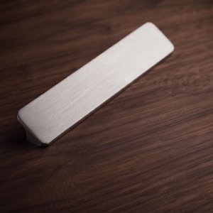 Puede incluir: Un tirador de mueble rectangular con acabado en metal plateado cepillado. El tirador presenta una superficie superior plana y alargada con esquinas ligeramente redondeadas y una base cónica que se conecta a los puntos de montaje.