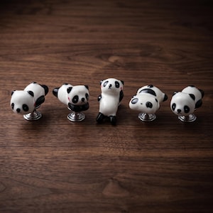 Puede incluir: Cinco pomos de cerámica para armarios con forma de osos panda en varias posturas. Cada panda es blanco con orejas, manchas en los ojos, extremidades negras y un pequeño detalle rosa en la mejilla. Montados sobre bases metálicas plateadas.