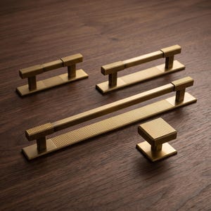 May include: A collection of four brass-toned cabinet hardware pieces, including three rectangular bar pulls and one square knob. Each piece features a textured, ribbed surface pattern on the handle and base, with a smooth, matte metallic finish.