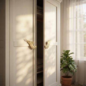 Puede incluir: Un armario alto de madera en color blanco roto con dos puertas ligeramente entreabiertas. Cada puerta cuenta con un tirador decorativo de metal dorado con forma de alas emplumadas, colocado horizontalmente en el centro de las puertas.