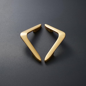 Puede incluir: Dos tiradores de armario de metal dorado con un acabado mate y suave. Cada tirador presenta un diseño angular en forma de V con extremos cónicos, colocados simétricamente para formar una figura de diamante. Herrajes de construcción sólida.