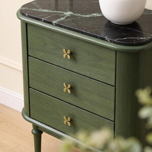 May include: A forest green wooden nightstand featuring three drawers with gold-toned, four-petal flower-shaped knobs. The top surface is made of black marble with white veining, and the unit stands on rounded legs with a smooth, curved frame.