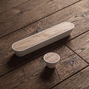 May include: A long, oval-shaped cabinet handle and a small, round cabinet knob. Both pieces feature a white base with a light brown wood grain inlay on the top surface, showcasing a clean, two-tone design for furniture hardware applications.
