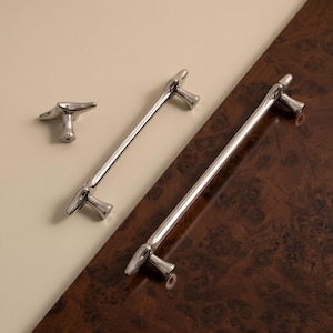 May include: A set of three polished silver-toned metal cabinet hardware pieces, including one T-shaped knob and two elongated bar pulls, each featuring a smooth, rounded design with tapered ends and cylindrical mounting posts for attachment.