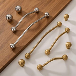 May include: A collection of cabinet hardware including two silver-toned curved handles with spherical ends, two silver-toned spherical knobs, three gold-toned curved handles with spherical ends, and three gold-toned spherical knobs arranged on surfaces.
