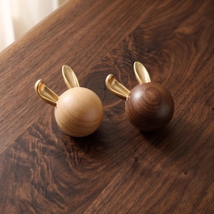 May include: Two spherical objects shaped like rabbit heads, each featuring two smooth gold-coloured metal ears. One sphere is crafted from light-toned wood with a visible grain, while the other is made from a darker, rich brown wood with a prominent grain.