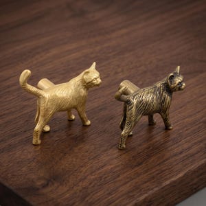 May include: Two cat-shaped cabinet knobs, one in a bright polished gold finish and the other in a darker, antiqued bronze finish. Each knob features a detailed feline figure standing with its tail raised, attached to a cylindrical base.
