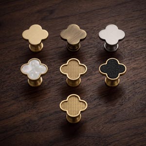 Könnte beinhalten: Sieben kleeblattförmige Schrankknöpfe in einer Gruppe angeordnet. Die Kollektion umfasst Ausführungen in massivem Messing, gebürstetem Gold, Silber, irisierendem weißem Perlmutt und schwarzer Textur, teilweise mit Gittermuster.