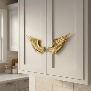 May include: A pair of gold-colored cabinet pulls shaped like detailed bird wings. Each pull features textured, layered feather patterns and is mounted horizontally on the center of two adjacent light gray kitchen cabinet doors.