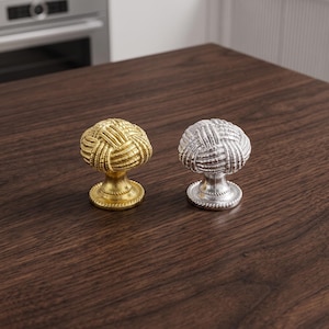 May include: Two cabinet knobs, one in a metallic gold finish and one in a metallic silver finish. Each knob features a spherical top with a textured, woven rope pattern and a circular base with a matching rope-like border detail.