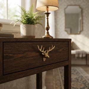 May include: A dark brown wooden console table featuring a single drawer with a metallic gold-toned deer head handle. The table surface holds a potted green fern in a white ceramic planter and a brass table lamp with a beige fabric shade.