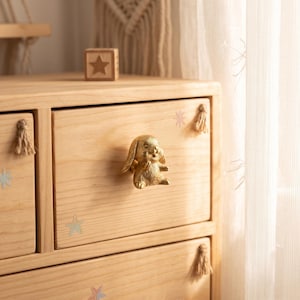 May include: A light-toned wooden chest of drawers featuring a metallic gold rabbit-shaped knob on the center drawer. The drawer front is decorated with small star decals and a beige tassel attached to the side. The wood grain is visible throughout.