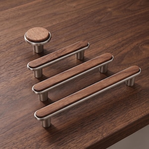 Puede incluir: Un conjunto de herrajes para muebles que consta de un pomo redondo y tres tiradores de cajón rectangulares. Cada pieza presenta una incrustación de madera marrón oscura en un marco de metal plateado cepillado con bordes redondeados.