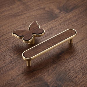 Peut inclure: Un bouton de meuble en forme de papillon et une longue poignée de tirage rectangulaire. Les deux pièces présentent une incrustation en bois brun foncé insérée dans un cadre en métal doré brossé, offrant un design bicolore pour la quincaillerie.