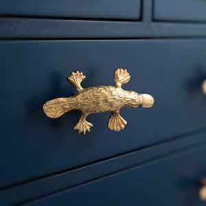 May include: A gold-colored metal drawer pull shaped like a platypus, featuring a textured surface and webbed feet, mounted on a smooth, dark navy blue drawer front. The handle is oriented horizontally with the bill pointing to the right side.