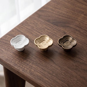 May include: Three cabinet knobs in a four-lobed flower shape with concentric circular grooves on the surface. The set includes one white knob, one light gold-toned metallic knob, and one dark bronze-toned metallic knob, each with a central stem.