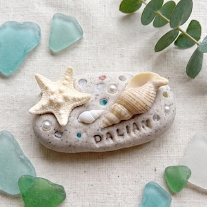 Puede incluir: Una piedra ovalada de color beige moteado con el nombre DALIAN, una estrella de mar blanca, una concha en espiral y pequeñas perlas incrustadas. La piedra está rodeada por varios trozos de vidrio marino liso en tonos azul claro, verde y blanco.