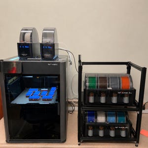 以下が含まれることがあります： ダークグレーの筐体と上部に2つのAMSユニットを備えた3Dプリンターで、ビルドプレート上に青い長方形の物体をプリントしています。その横には黒い金属製のラックがあり、それぞれ4つのフィラメントスプールが入った2つのAMSユニットが設置されています。