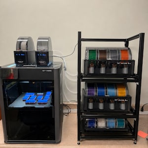 Może przedstawiać: Drukarka 3D z ciemnoszarą obudową i dwiema jednostkami AMS na górze, drukująca niebieskie prostokątne obiekty. Obok znajduje się czarny metalowy regał z trzema ułożonymi w stos jednostkami AMS, z których każda zawiera cztery szpule filamentu.