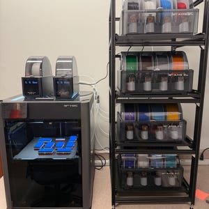 May include: A Bambu Lab 3D printer with two AMS units on top, printing small rectangular objects on the build plate. Beside it stands a tall black metal shelving unit holding four additional AMS units, each containing multiple colorful 3D printing filament spools.
