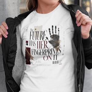 Puede incluir: Una camiseta blanca de manga corta con un diseño gráfico en negro y rojo oscuro. El texto dice, The FUTURE HAS HER FINGERPRINTS ON IT, acompañado de una ilustración detallada de una huella dactilar negra y varias formas geométricas.
