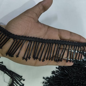 Puede incluir: Un fleco de cuentas negras con largas cuentas cilíndricas negras que cuelgan de una cinta de tela tejida. Las cuentas están dispuestas en una fila uniforme, cada una terminando en un pequeño nudo redondeado, creando un borde decorativo.