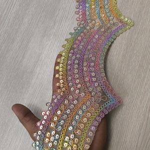 May include: A long, decorative trim featuring a sheer mesh base with an intricate, wavy embroidered pattern. The embroidery consists of vibrant, multi-colored thread in shades of yellow, orange, pink, purple, and blue, accented with small, shiny sequins.