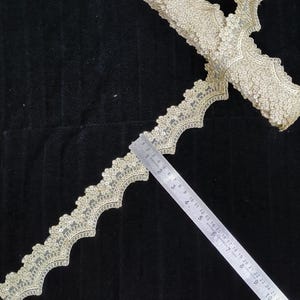 Peut inclure: Une bordure en dentelle dorée avec un bord festonné et une broderie complexe ornée de petites paillettes. Une règle en métal est placée le long de la bordure, indiquant une largeur d'environ 4,5 cm pour le motif décoratif en dentelle.
