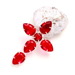 May include: A cross-shaped pendant featuring five vibrant red faceted gemstones set in silver-toned metal. The design includes four pear-shaped stones arranged in a cross pattern with a single round stone at the centre and a silver bail at the top.