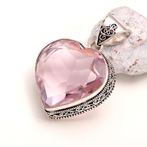 May include: A heart-shaped pendant featuring a faceted light pink gemstone set in a decorative silver frame with intricate filigree detailing along the edges and a matching silver bail at the top for attaching to a necklace chain.