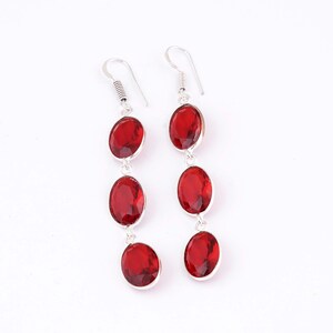 May include: A pair of drop earrings featuring three vertically aligned, oval-shaped red faceted stones set in silver-toned metal. Each earring is suspended from a simple silver-toned hook wire, creating a linear design with a polished finish.