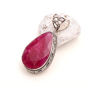 May include: A teardrop-shaped pendant featuring a faceted, deep pink gemstone set in a decorative silver-toned metal frame with intricate filigree detailing along the edges and a large, ornate bail at the top for attaching to a necklace chain.