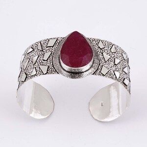 Puede incluir: Un brazalete de plata con una piedra preciosa de color rosa oscuro en forma de lágrima en el centro. La banda tiene un acabado texturizado y oxidado con formas geométricas de plata incrustadas. El interior es liso y lleva el sello 925.