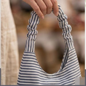 May include: A hobo-style handbag featuring a blue and white horizontal striped pattern. The bag includes a single gathered, scrunchie-style handle that matches the striped fabric of the main body, creating a cohesive and uniform appearance throughout.