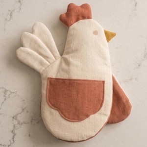 May include: A chicken-shaped oven mitt in off-white fabric with a rust-colored comb, a matching rust-colored wing-shaped pocket, and a mustard yellow beak. The mitt features a three-part tail design and a simple circular eye detail on the side.