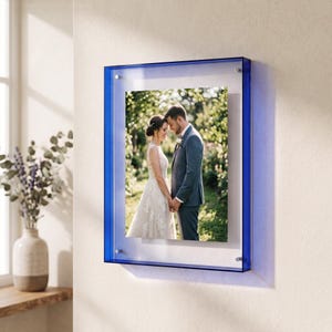 May include: A rectangular wall-mounted picture frame featuring a vibrant blue acrylic border. The frame holds a photograph of a couple standing together in a garden setting, with the frame secured by four small silver-toned metal mounting bolts.