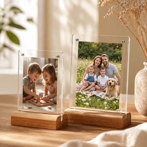 May include: Two vertical clear acrylic photo frames with light-toned wooden bases. Each frame features silver-colored metal hardware at the top corners to secure the acrylic panels, holding photographs of children and a family with a dog.