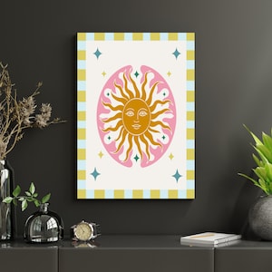 Puede incluir: Una impresión de arte mural rectangular que presenta un sol dorado central con un rostro humano, rodeado por una forma ovalada rosa. El fondo es blanco hueso con estrellas dispersas en color verde azulado y amarillo. Un borde a cuadros amarillo y azul claro enmarca la impresión.