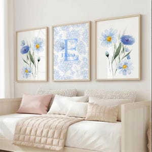 May include: A set of three framed wall art prints featuring light blue and white floral illustrations. The center print displays a large letter E with the name Eliana printed below it, surrounded by a delicate blue floral pattern on a white background.
