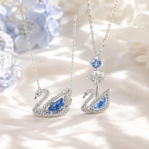 Puede incluir: Dos collares plateados con colgantes en forma de cisne. Un colgante es un cisne incrustado con cristales transparentes y una piedra azul central. El segundo colgante presenta un cisne coronado por dos elementos de cristal geométricos.