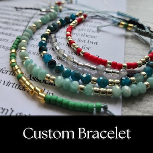 May include: A collection of five layered bracelets featuring various beads including faceted green, blue, and clear stones, matte red beads, and metallic gold cylindrical spacers. The bracelets are arranged in a circular pattern on a white card.