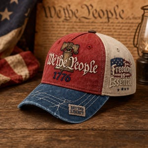 Può includere: Un cappellino da baseball dall'aspetto vissuto con pannello frontale rosso ricamato con la Liberty Bell, il testo We the People e 1776. Il pannello laterale è beige con una grafica a bandiera e il testo Freedom is Essential. La visiera blu ha cuciture bianche.